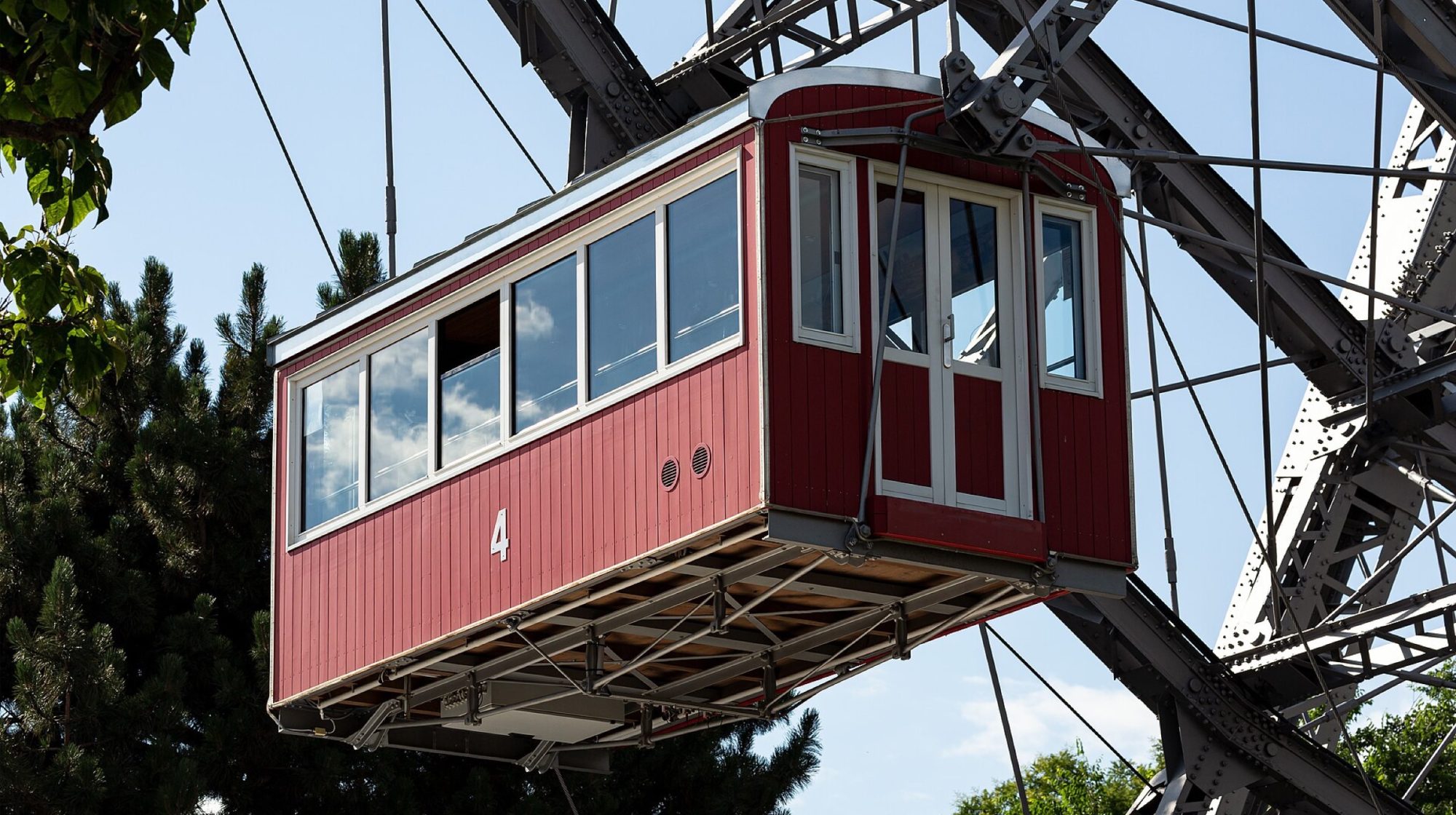 A red wooden gondola of the Wiener Riesenrad against a blue sky, with the wheel's steel structure behind it